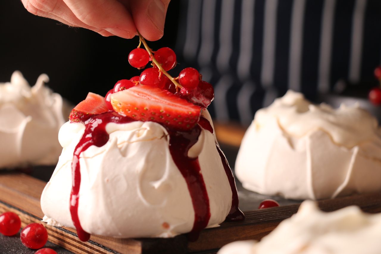 Crop anonymous cook decorating tasty crunchy meringue cake with ripe strawberry slices and sprig of colorful red currants on top of sweet red sauce in restaurant kitchen